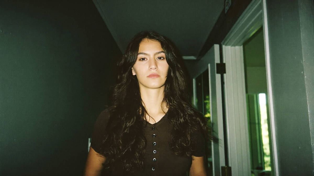 A young woman standing in a dark green hallway beside her open front door, lit by hard direct flash