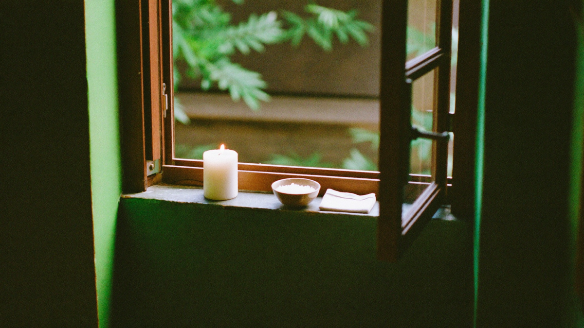 White pillar candle, a bowl of coarse salt and folded linen on the sill of an open window in a green room
