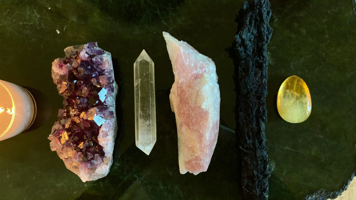 Five crystals laid side by side on dark green stone in candlelight: amethyst cluster, clear quartz point, rose quartz, black tourmaline rod and citrine