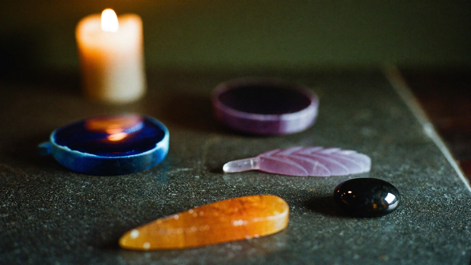 Flat polished palm stones in blue, purple, rose and amber laid on dark stone beside a burning candle