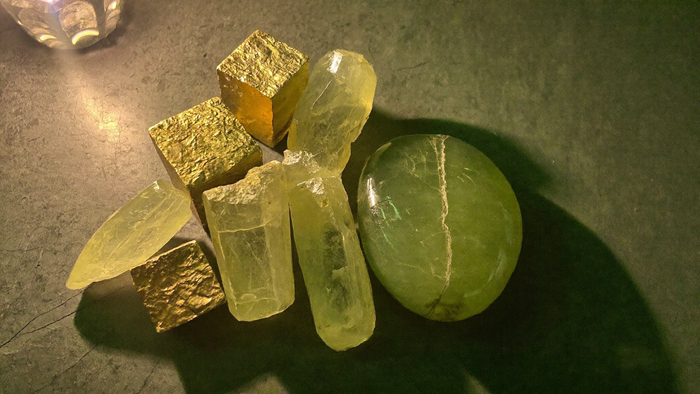 Golden pyrite cubes, pale citrine points and a green aventurine pebble on dark stone in candlelight