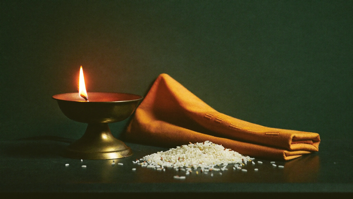 Lit brass oil lamp beside folded saffron cloth and a scattering of rice grains on dark stone