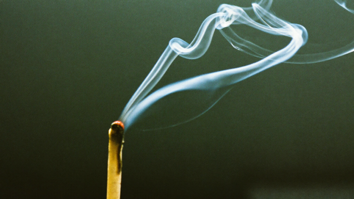 Burnt-out matchstick with a thin ribbon of smoke curling upward against a dark green wall