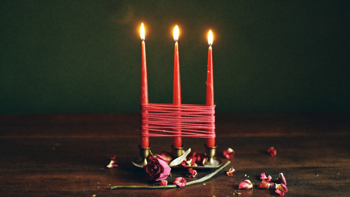 Three red taper candles bound together with red thread beside a dried rose and scattered petals on a dark wooden table
