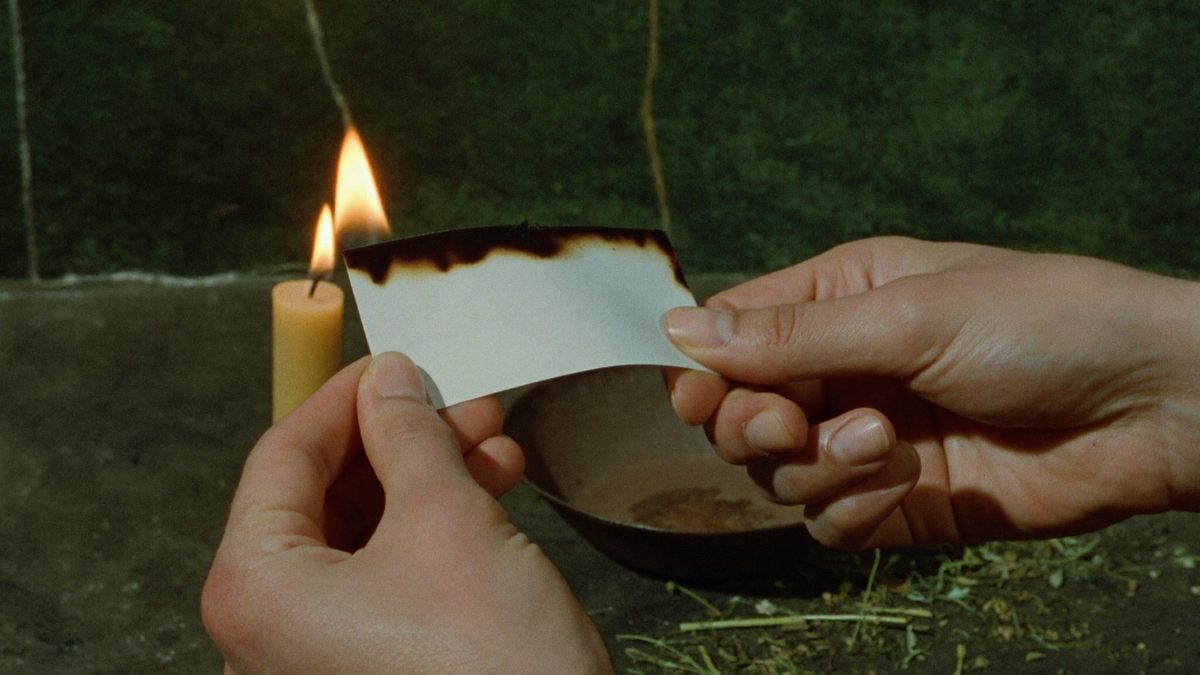 Two hands holding a charred slip of paper above an iron bowl beside a lit candle and scattered dried herbs
