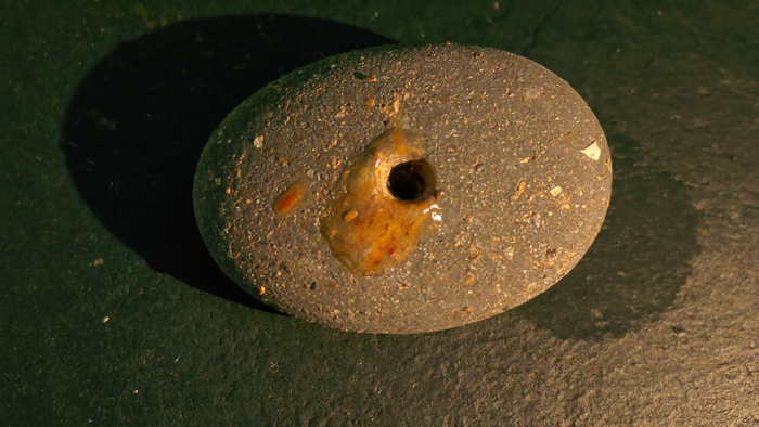 Wet grey beach pebble with a natural hole worn through it, lit by candlelight on dark slate