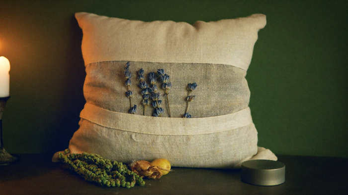 A linen herb pillow with lavender sprigs, hop cones and a small tin beside a lit candle on a dark table