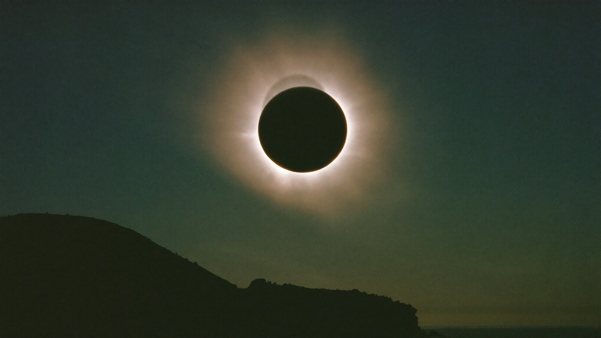 Total solar eclipse with the black disc of the moon and a glowing white corona above a dark coastal hill at dusk