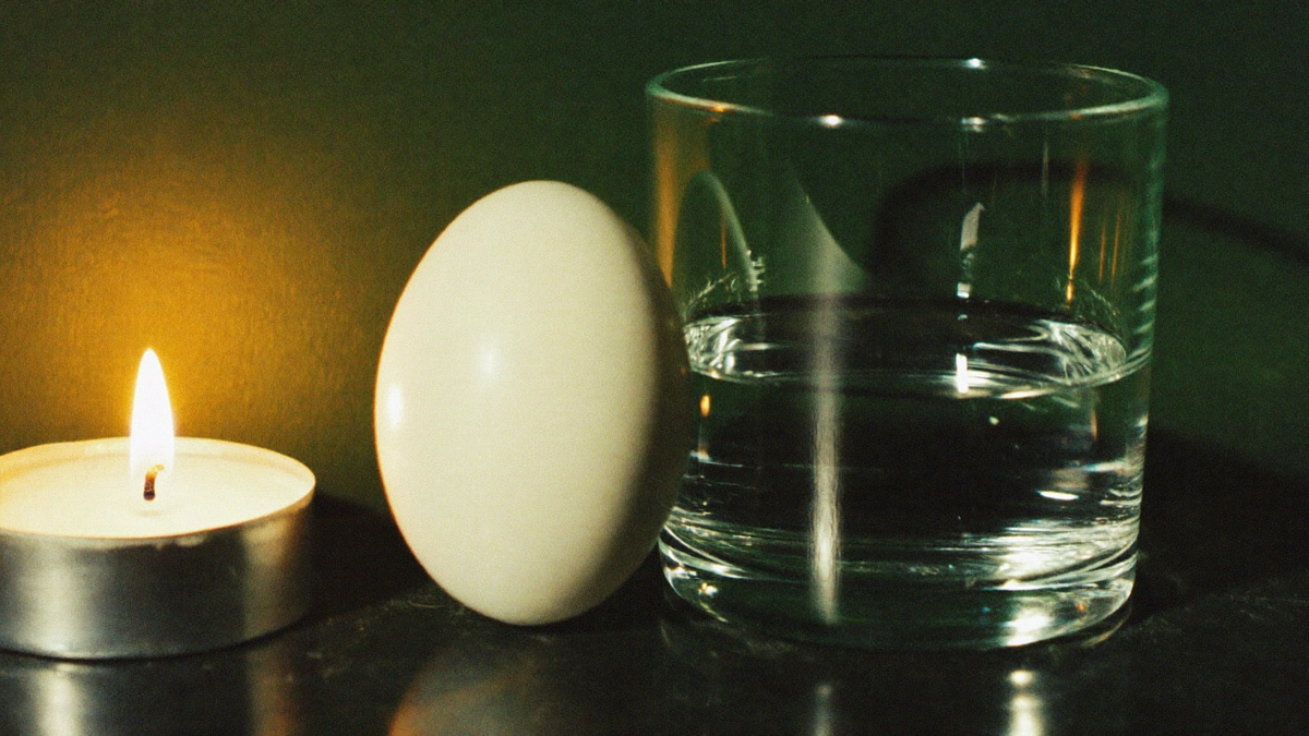 A single white egg beside a clear glass of water on dark stone, lit by one candle