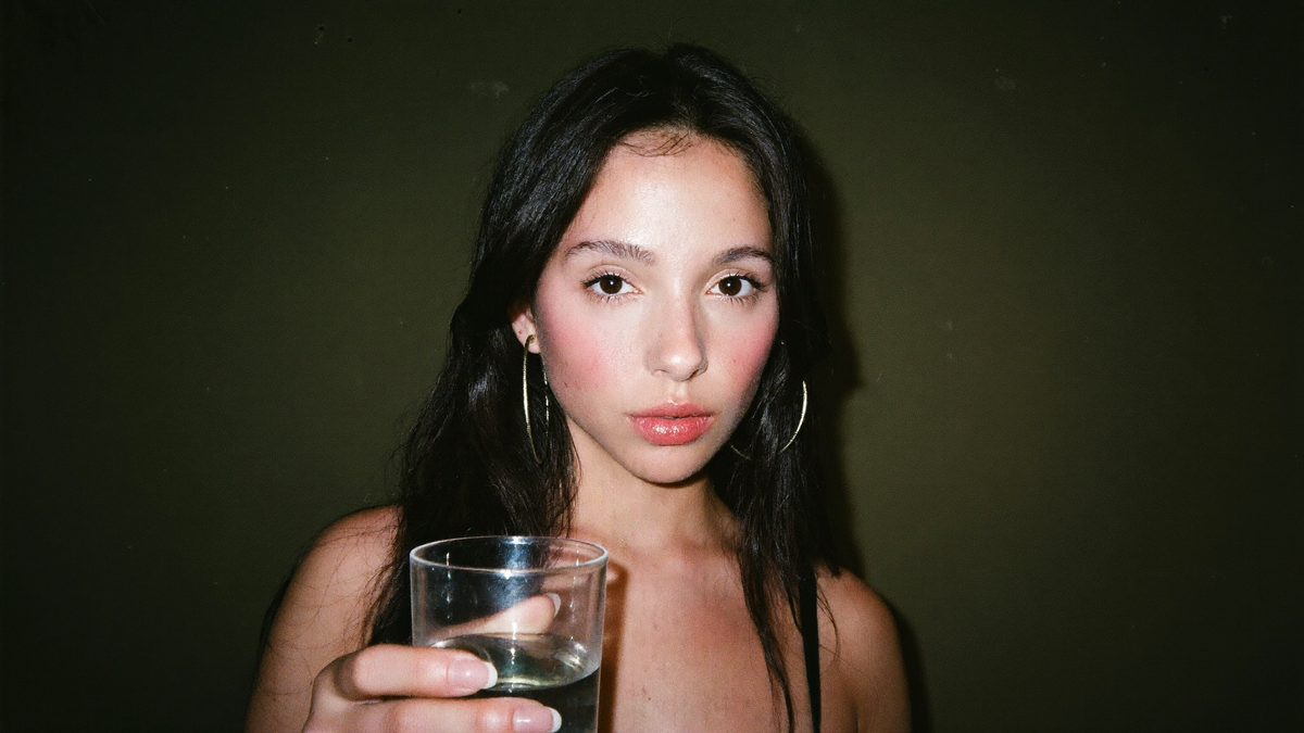 Young woman with dark hair holding a plain glass of water, direct flash, olive green wall behind