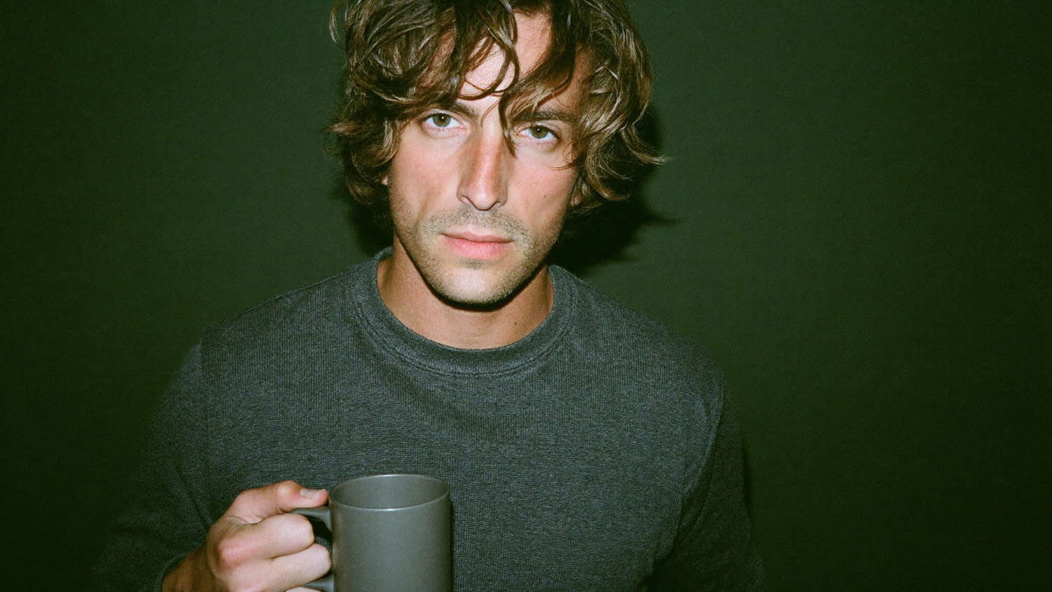 Young man holding a plain grey mug against a dark green wall, lit by direct flash
