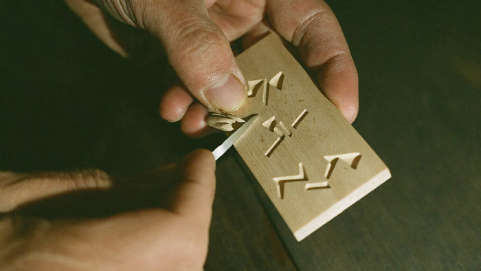 Weathered hands carving angular rune shapes into a pale strip of ash wood with a small knife in warm light