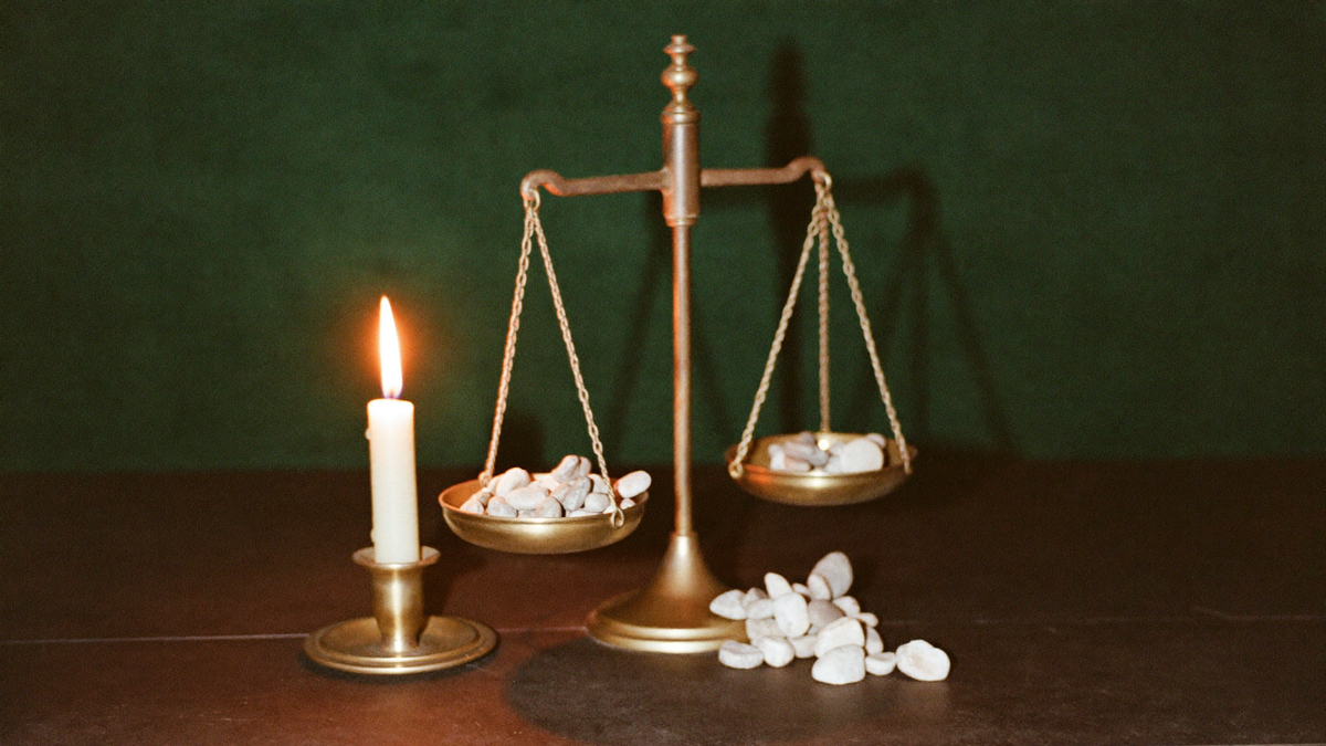 A small brass balance with white counting pebbles in both pans, lit by one candle against dark green