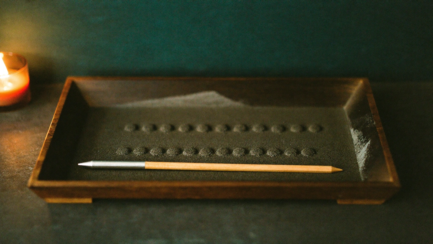 Rows of small round dots pressed into fine dark sand in a shallow wooden tray, a pointed stylus resting across it