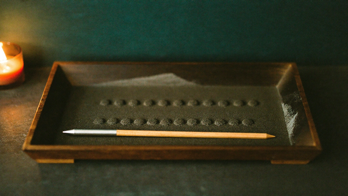 Rows of small round dots pressed into fine dark sand in a shallow wooden tray, a pointed stylus resting across it