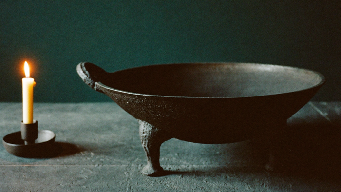 Antique three-legged cast iron cauldron on dark stone beside a single burning candle against a dark green wall