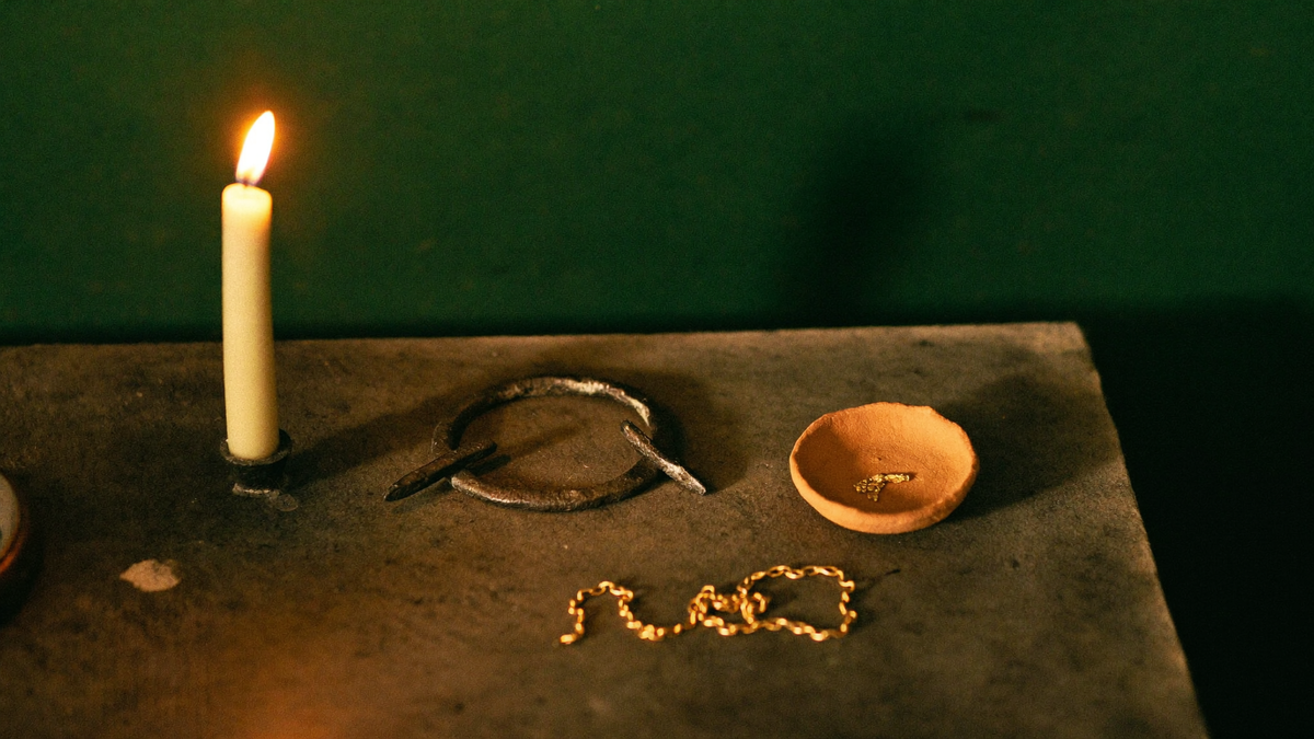 An iron ring, a clay crucible holding a trace of gold and a fine gold chain on dark stone by candlelight