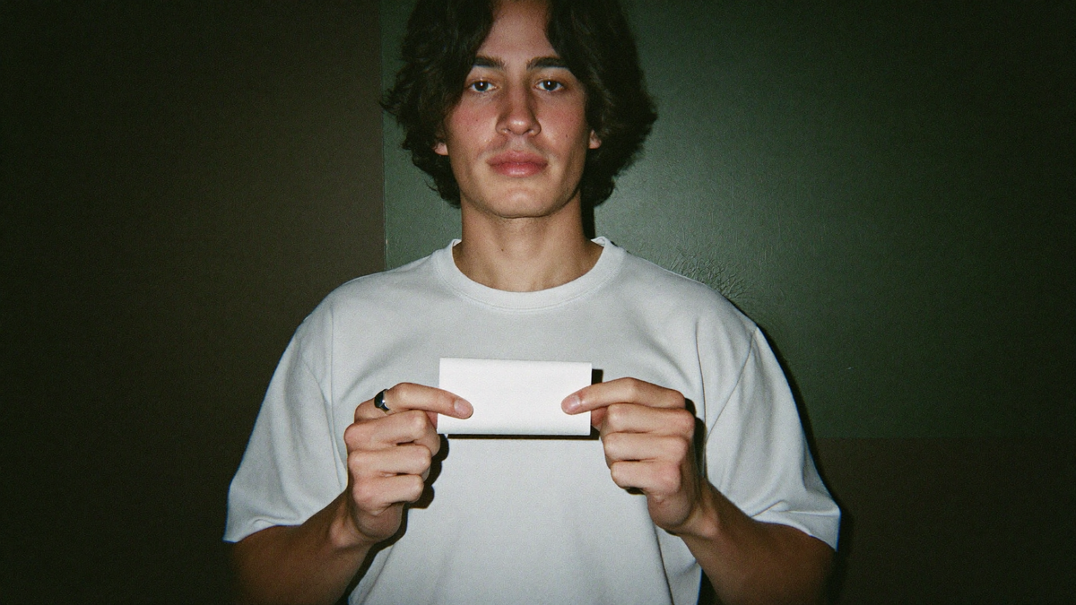 Young man holding a blank white paper slip in both hands against a dark green wall
