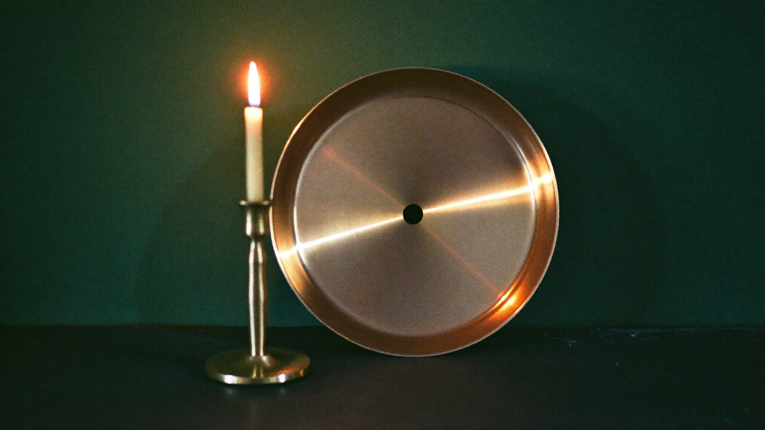 A polished brass sun disc beside a lit taper candle on dark stone against a deep green wall