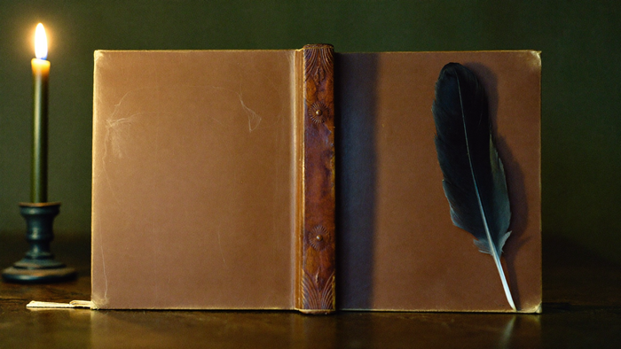 Old leather-bound book standing open with a black quill feather and a lit candle beside it