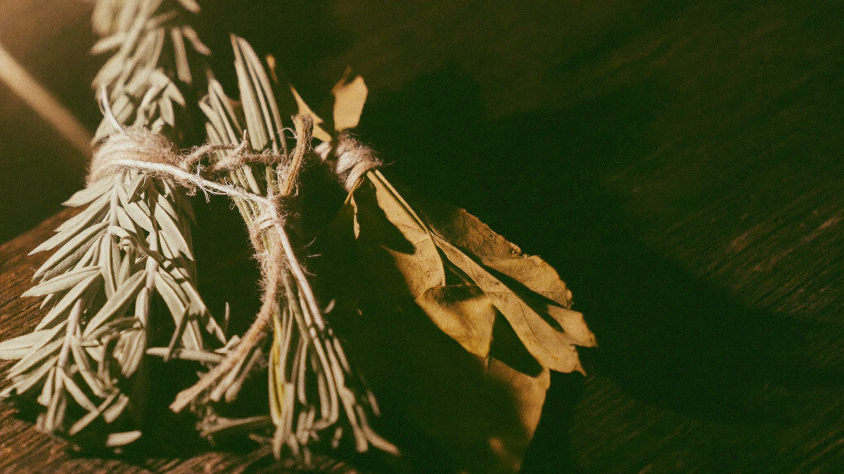 Bundles of dried rosemary, mugwort and bay leaves tied with twine on dark wood in warm candlelight