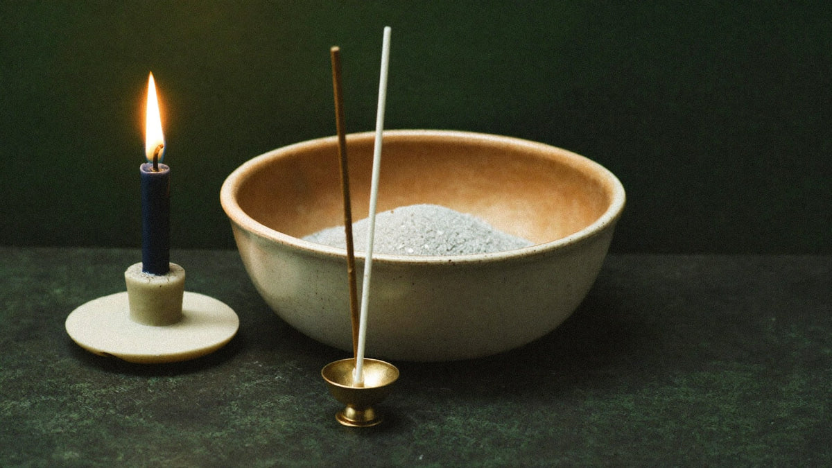 A ceramic bowl of ash, a brass stick holder and a lit candle on a dark stone surface