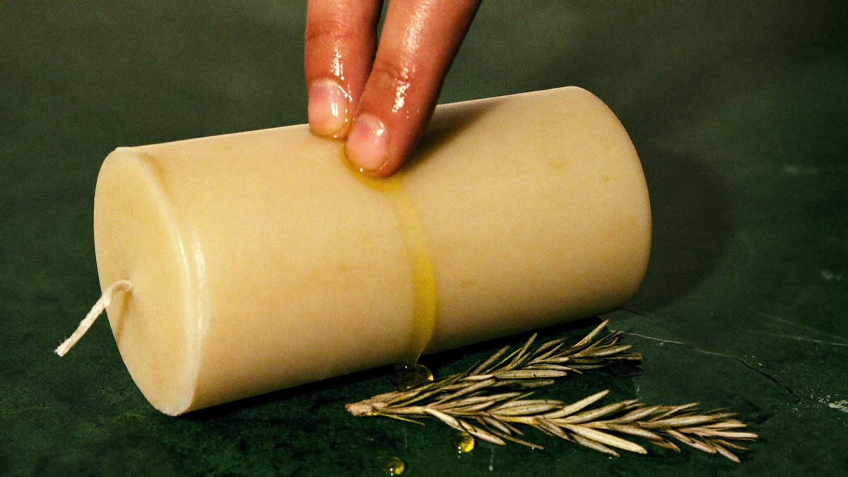 Oiled fingertips drawing a line of ritual oil down a cream pillar candle lying on dark stone beside dried rosemary