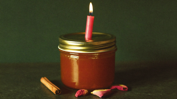 Closed jar of amber honey with a lit pink candle on its brass lid, cinnamon stick and rose petals on dark stone