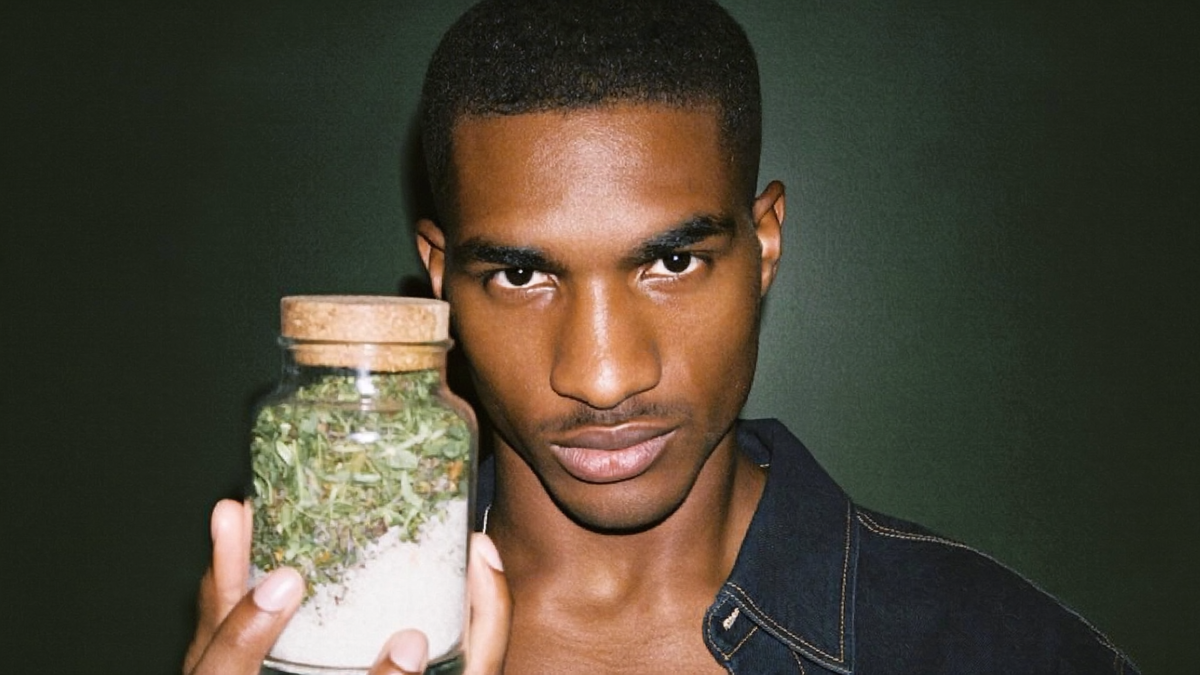 Young man holding a small corked glass jar packed with layers of dried herbs above coarse salt