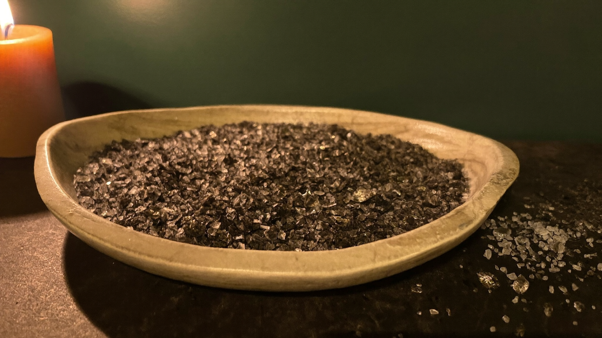 Coarse black ritual salt heaped in a pale stone dish beside a lit candle, spilled crystals on dark stone