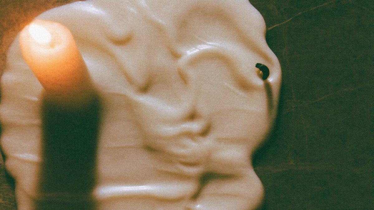 Pool of cooled pale candle wax set into ridges and folds on a dark surface beside a burning candle