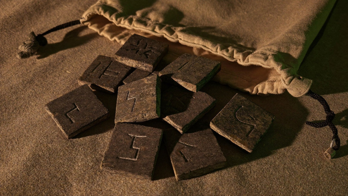 Carved stone rune tiles spilling from a canvas drawstring bag onto coarse linen in warm candlelight
