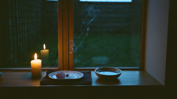 Small home altar on a wooden windowsill at dusk with a burning candle, a smoking incense stick in a ceramic dish and a bowl of water