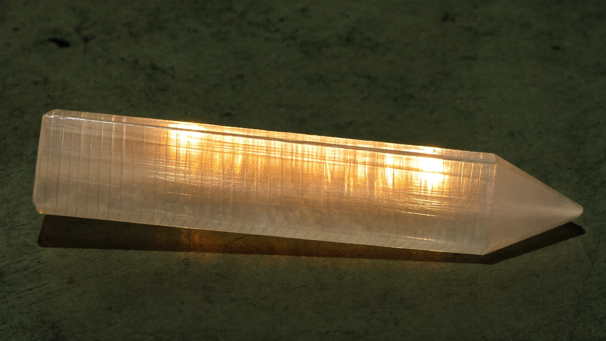 A polished satin spar selenite wand lit from the side, its fine parallel fibres glowing on dark stone