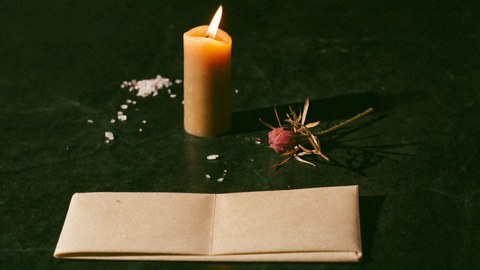 Folded petition paper on a dark stone surface with a lit candle, scattered salt and a dried sprig behind it