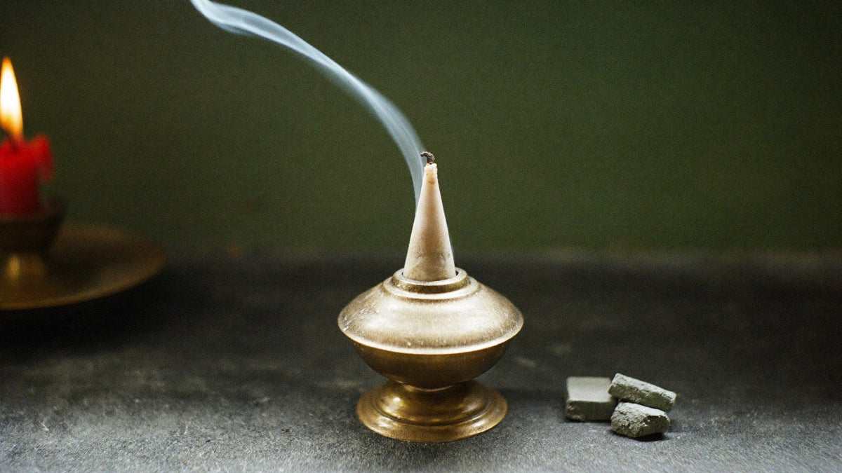 A brass cone incense burner with a thin ribbon of smoke rising beside two charcoal blocks