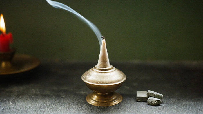 A brass cone incense burner with a thin ribbon of smoke rising beside two charcoal blocks