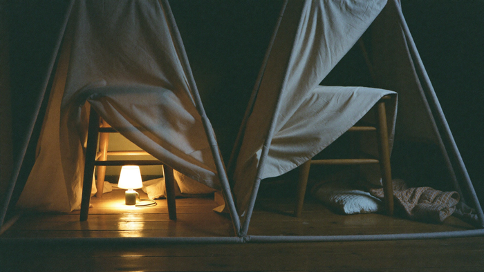 A blanket fort built over two wooden chairs with a small lamp glowing inside on bare floorboards