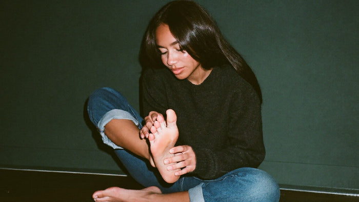 Woman in a dark sweater sits cross-legged and holds the bare sole of her itchy foot against a green wall