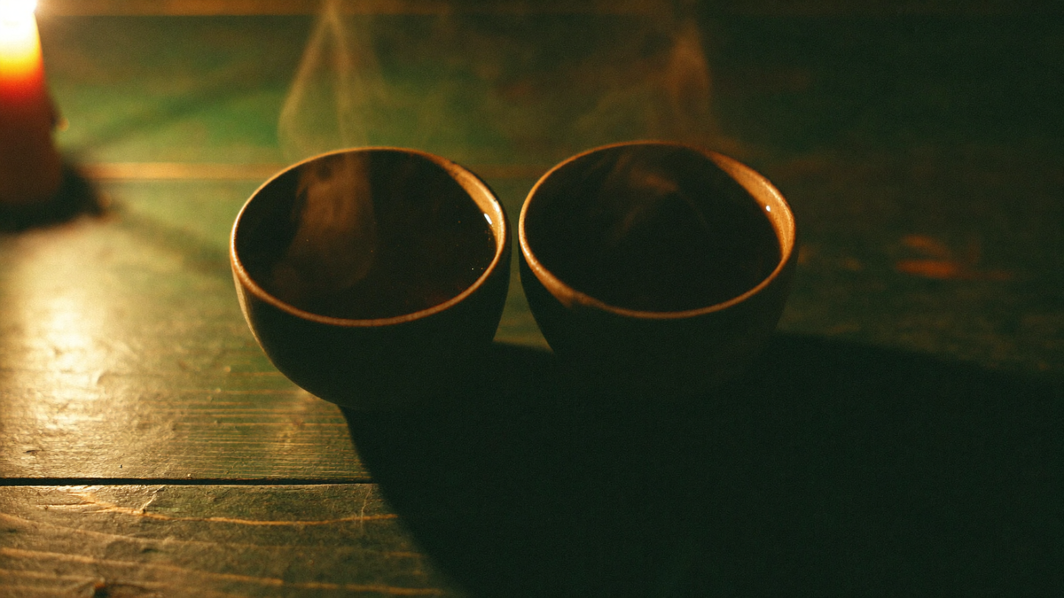 Two dark ceramic cups of tea facing each other on a worn green table in candlelight