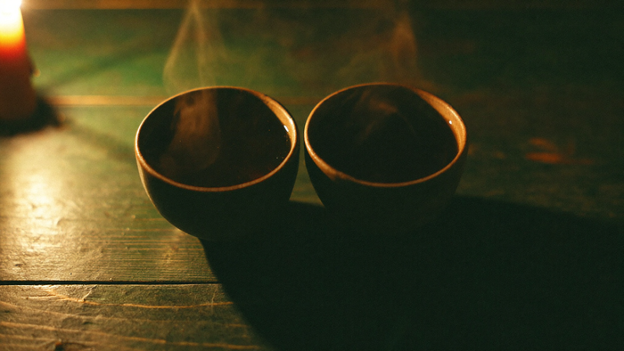 Two dark ceramic cups of tea facing each other on a worn green table in candlelight