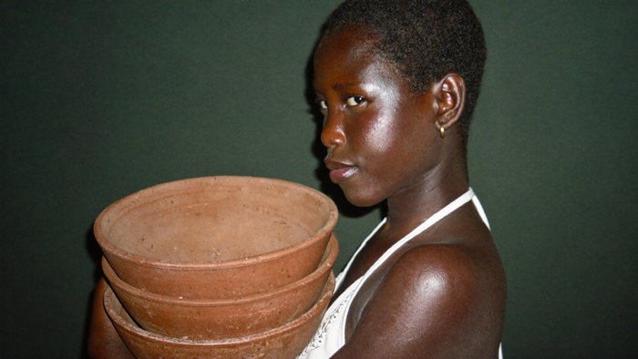 A young woman carrying a stack of plain clay bowls against a dark green wall, caught mid-task