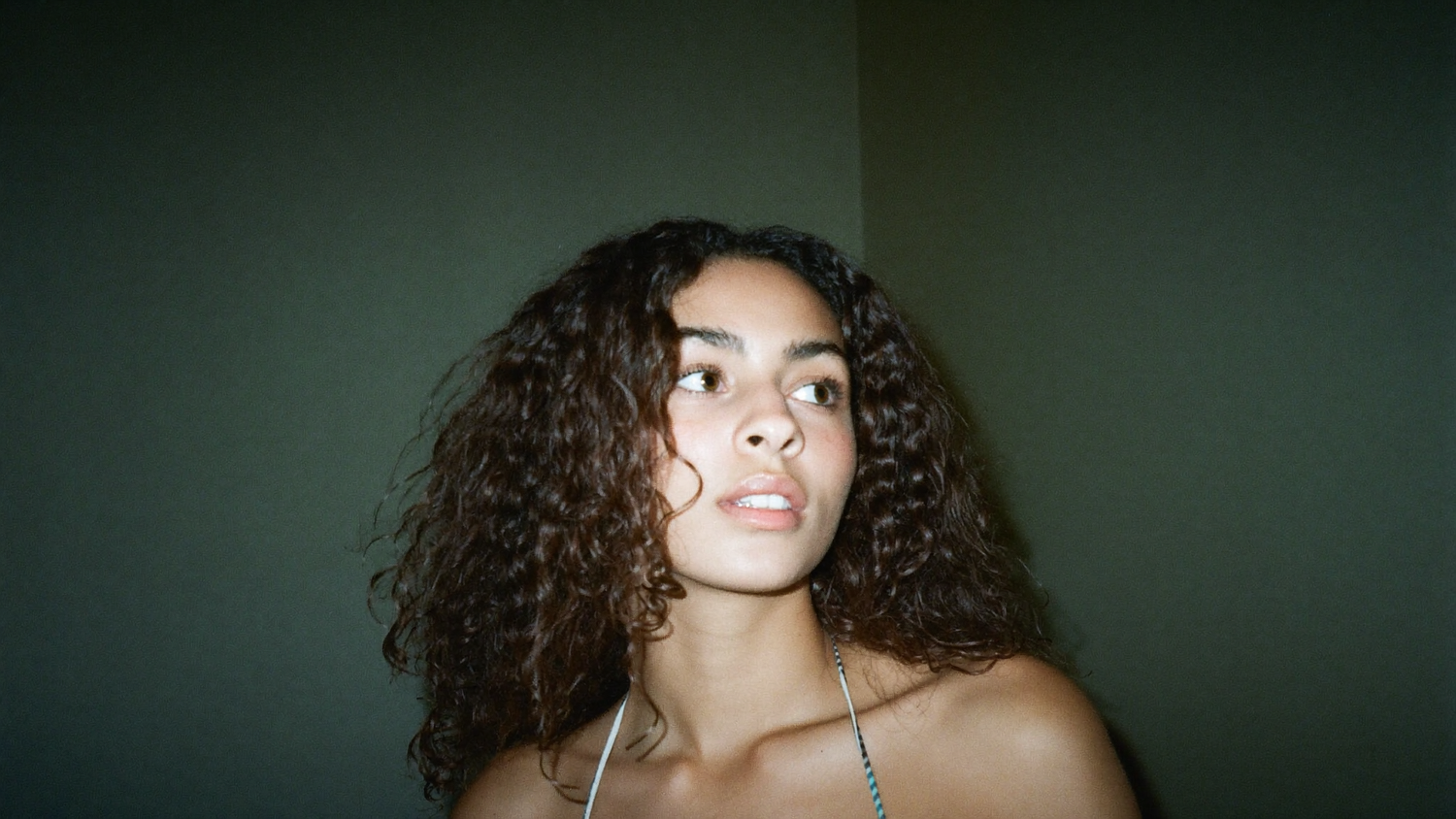 A young woman with dark curly hair photographed against a muted dark green wall, looking past the camera