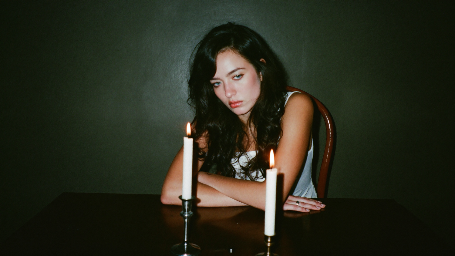 A woman sitting alone at a dark table behind two burning candles, lit by hard direct flash.