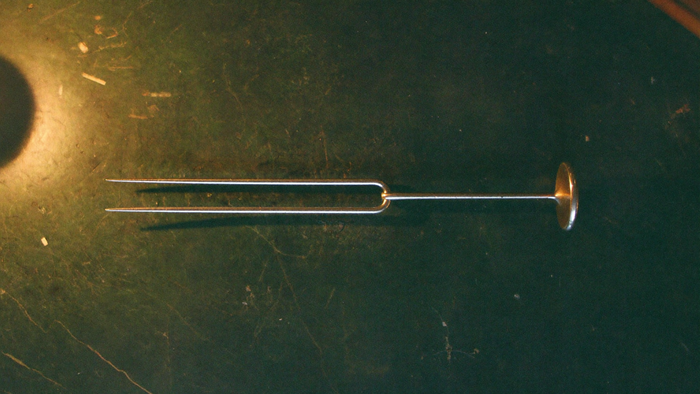 A steel tuning fork lying flat on dark stone in warm candlelight, both prongs clearly visible