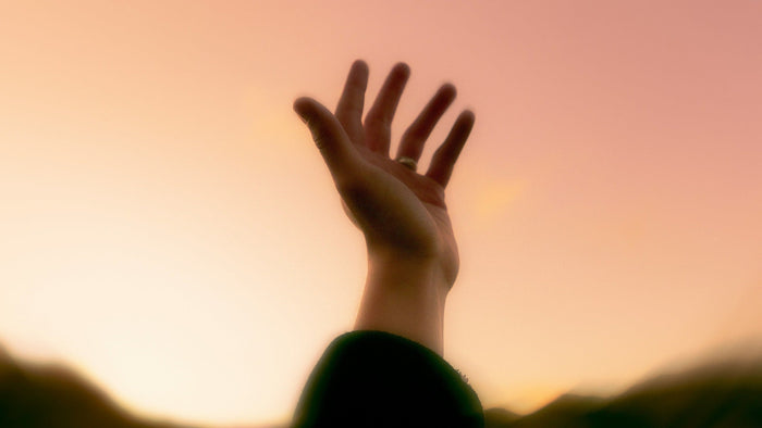 An open hand raised toward a soft rose and gold evening sky above dark hills, a gesture of prayer and remembrance