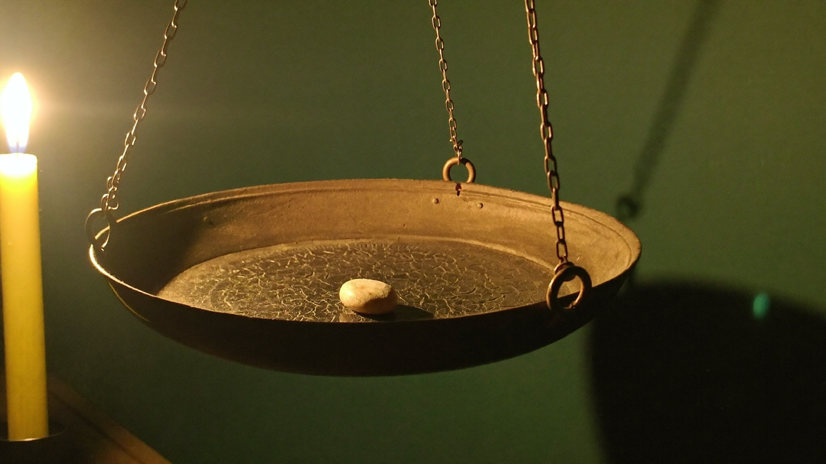 Brass pan of an old balance scale hanging on chains with a single pale stone, lit by a candle