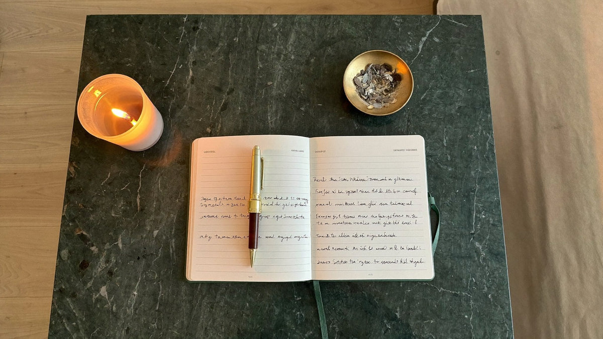 An open dream journal with a pen beside a lit candle and a small brass bowl of dried herbs