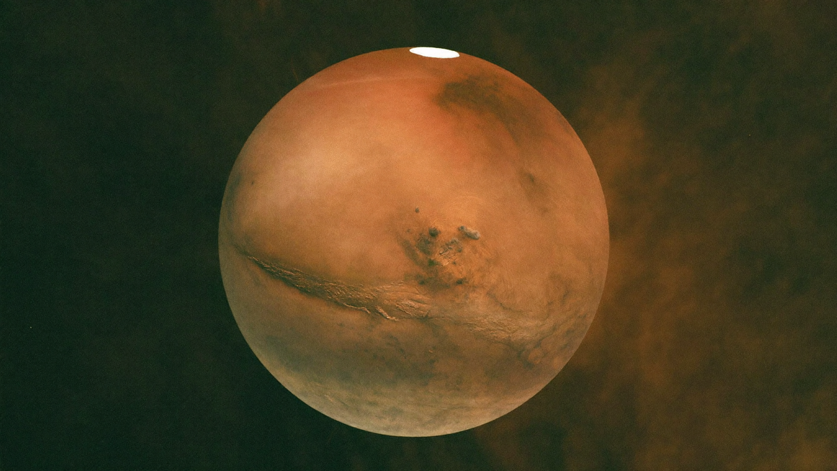 Mars as a rust-red globe with a bright white polar cap and a long dark canyon running across its middle against a dark sky