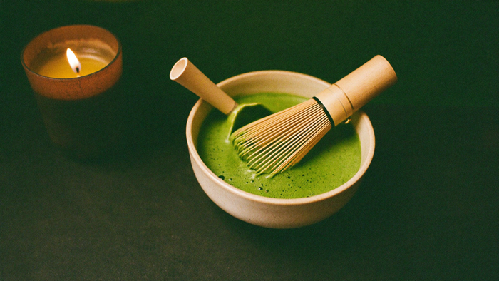 Bowl of whisked matcha with a bamboo whisk resting in the green foam beside a lit candle on a dark surface